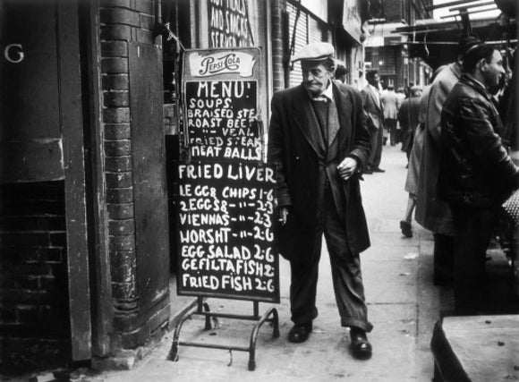 A man reads a chalk board on the street outside a cafe:1961